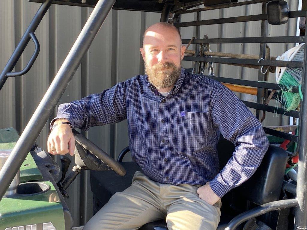 A bearded man, Greg Wilson, sits in a farm vehicle and smiles at the camera.