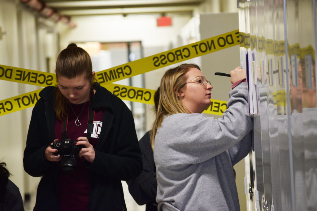 A girl holding a camera looking at pictures and the girl next to her taking notes by leaning on wall. All this with "crime do not cross" tape behind them.