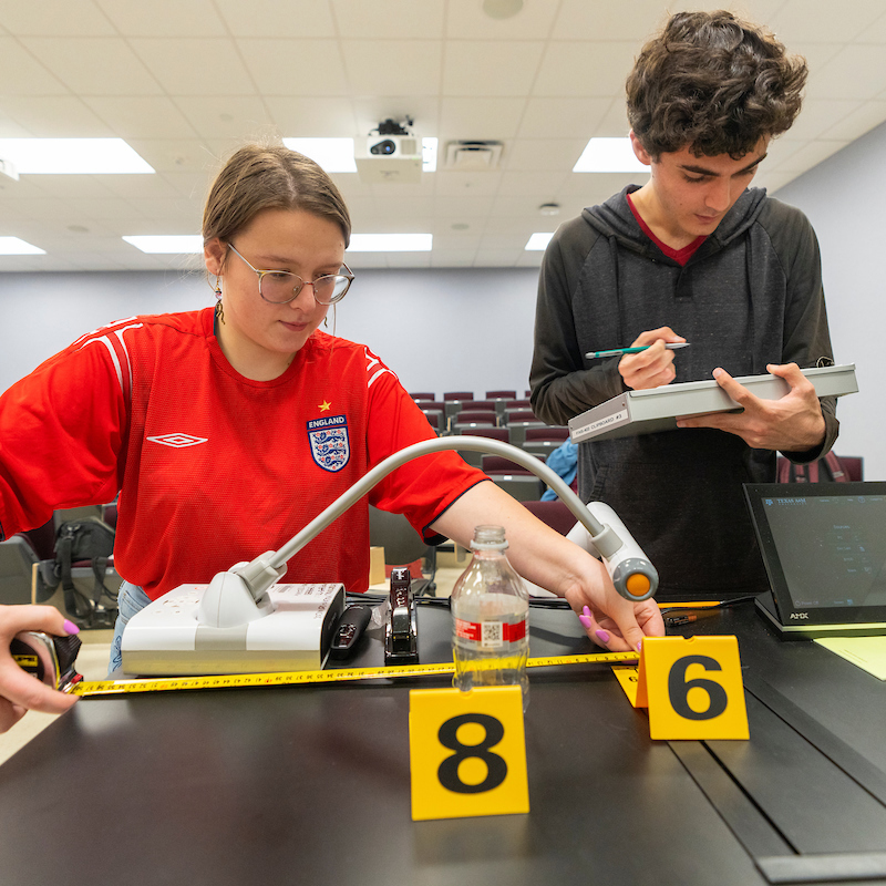 One student measures forensic evidence with a tape measure while another writes on a clipboard.